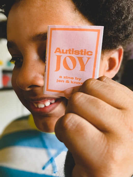 Smiling Black child holding up miniature zine cover that says Autistic Joy