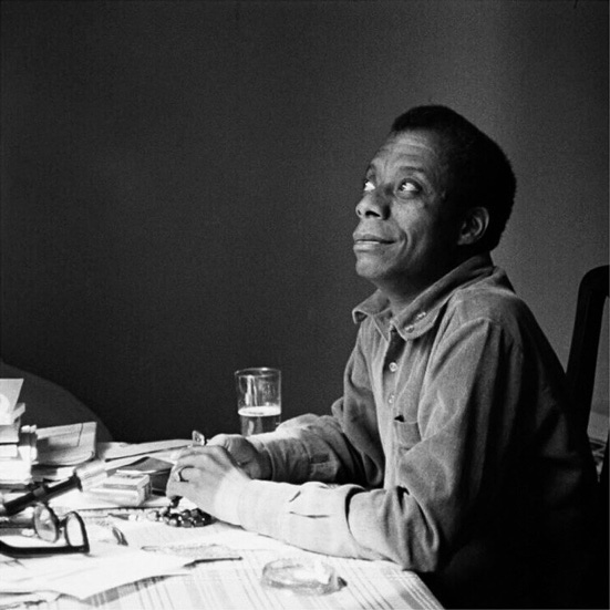 Black and white photo of James Baldwin sitting at a kitchen table looking up.