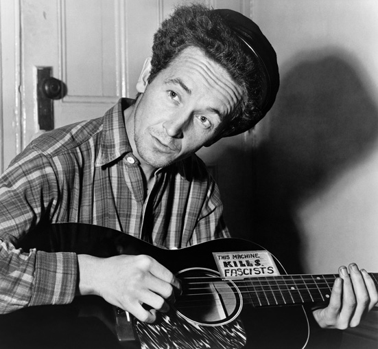 A black and white photo of Woody Guthrie playing a guitar with the message "This machine kills fascists".