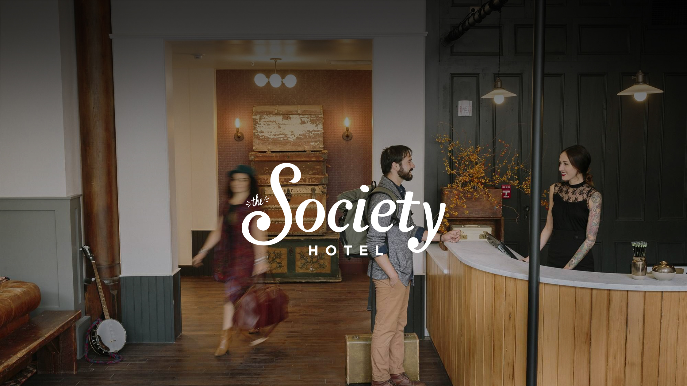 A photo of The Society Hotel's front desk. A patron casually stands smiling at the hotel clerk, and she returns the smile. A guest is blurred in the background indicating a brisk walk through the lobby.