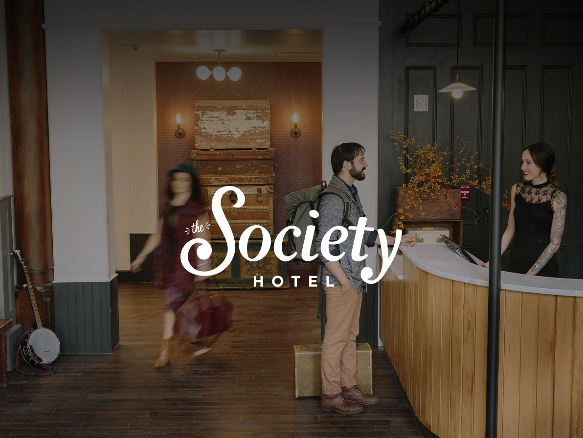 A photo of The Society Hotel's front desk. A patron casually stands smiling at the hotel clerk, and she returns the smile. A guest is blurred in the background indicating a brisk walk through the lobby.