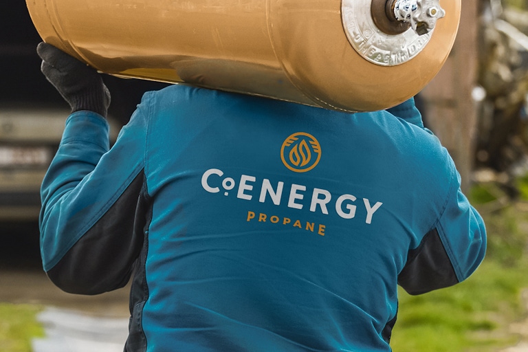 a photograph of a person carrying a propane tank, their back to the camera with the CoEnergy propane logo on the back of their jacket.