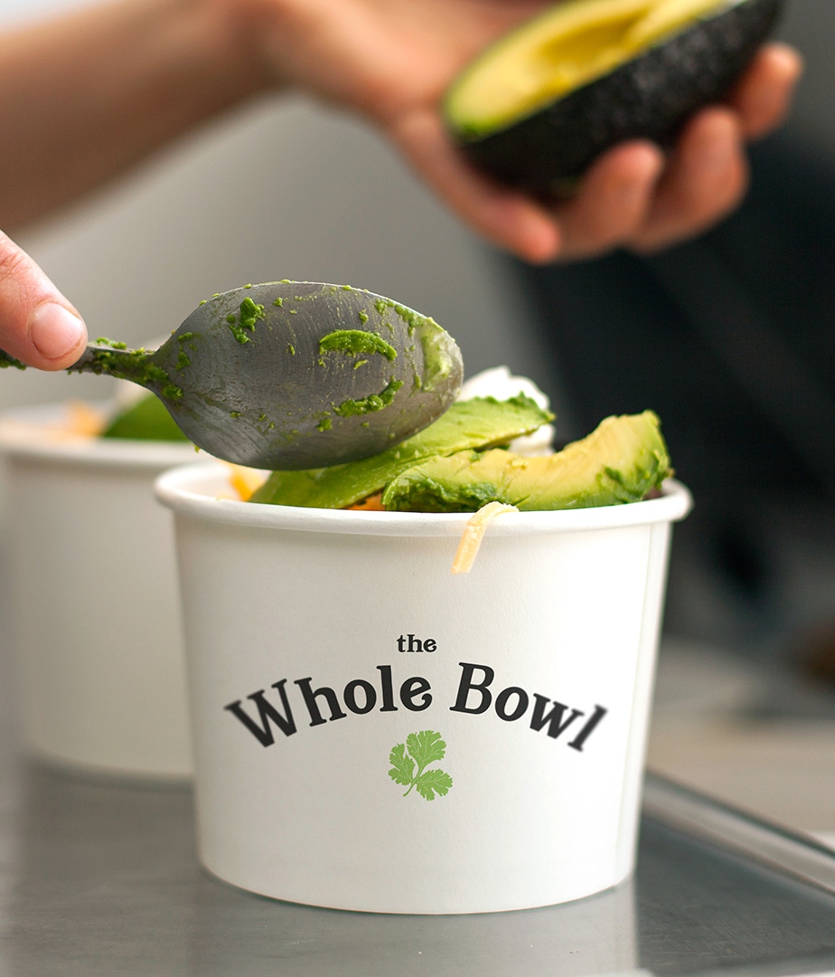 A photograph of someone scooping fresh avocado into a Whole Bowl with the logo on the cup.