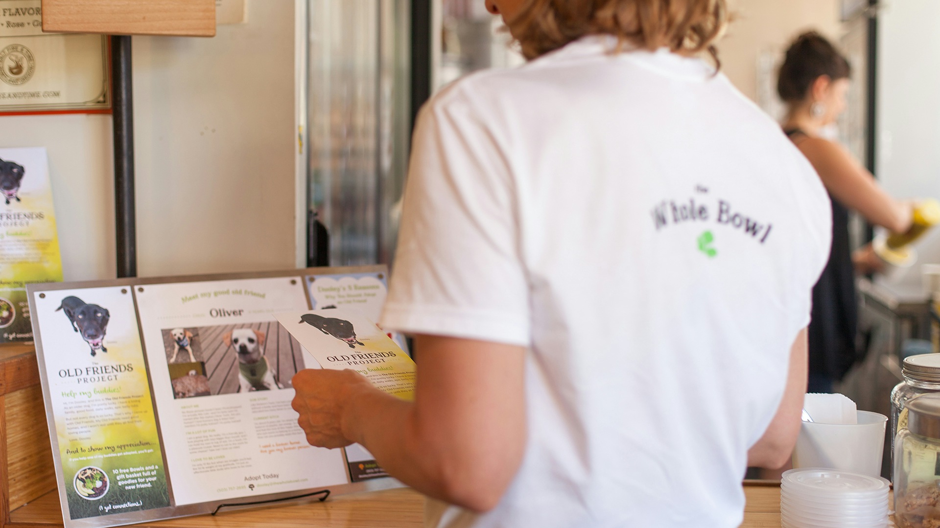 A photograph of a woman standing in a Whole Bowl restaurant wearing a white shirt with the Whole Bowl logo.