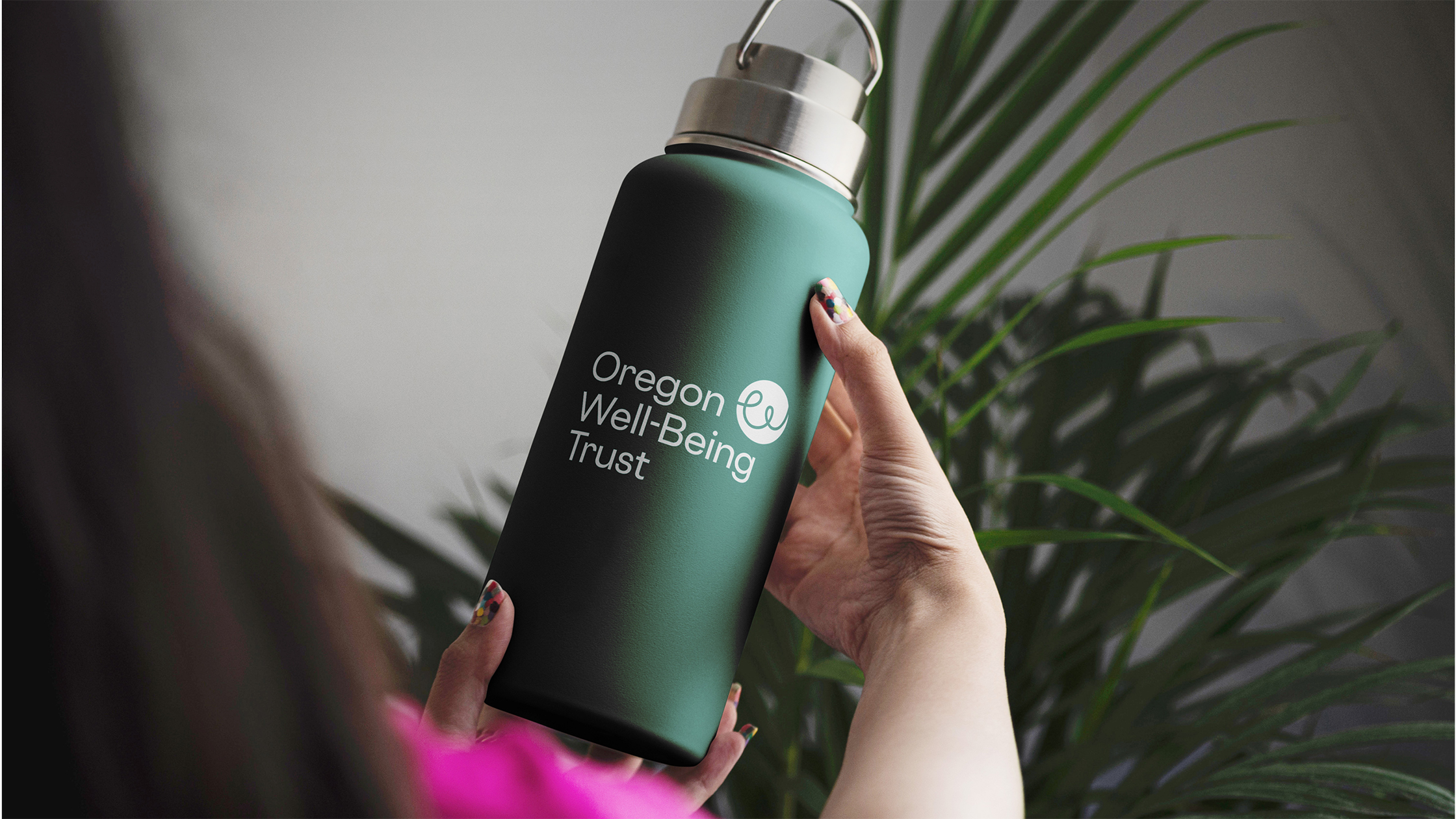 A close-up photograph of a hand holding a dark green water bottle with the Oregon Well-Being Trust's new logo printed on the front.