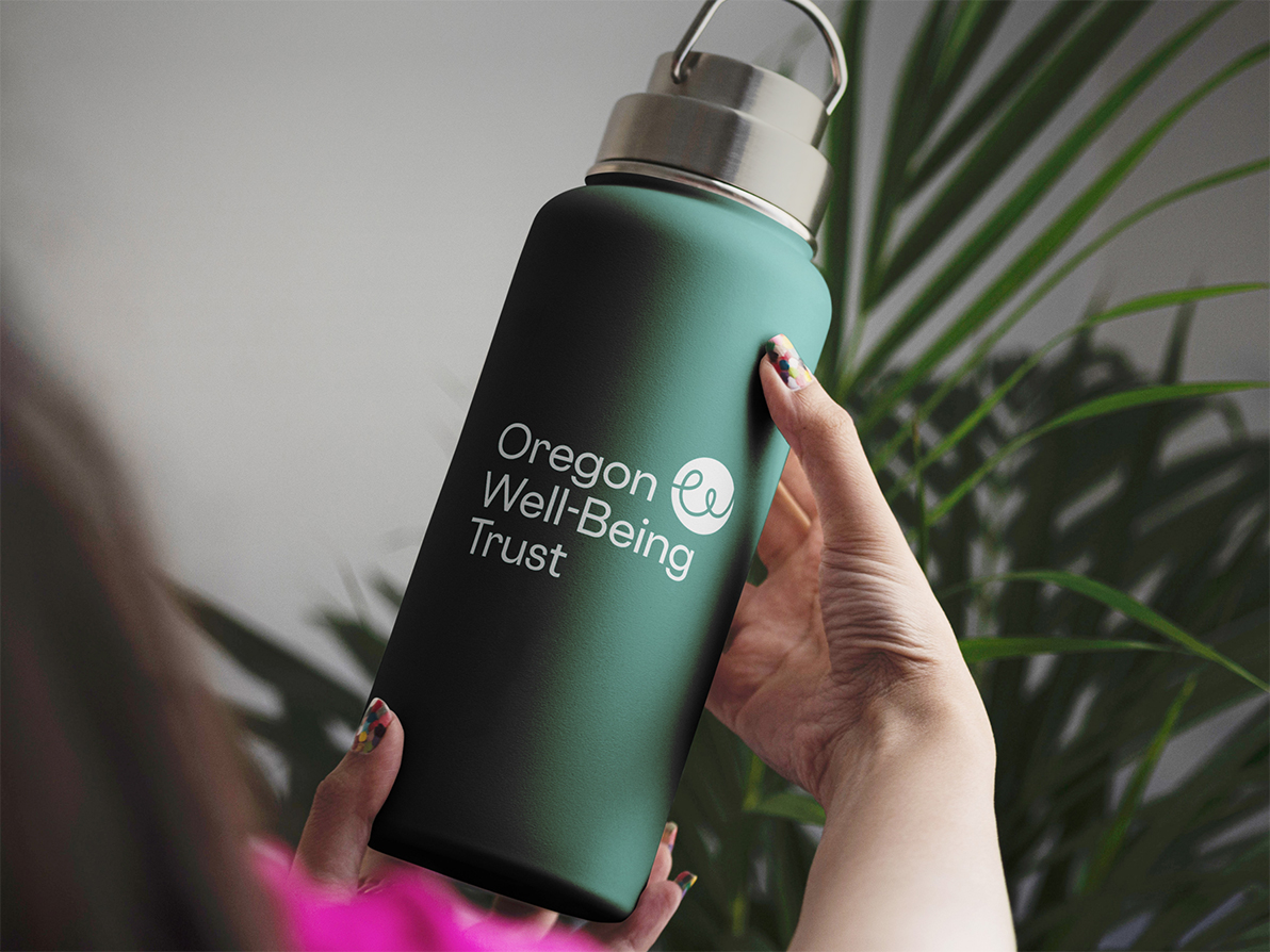 A close-up photograph of a hand holding a dark green water bottle with the Oregon Well-Being Trust's new logo printed on the front.